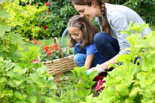 A woman and a young girl are gardening together in a lush outdoor space, crouching amongst vibrant green foliage and flowering plants. The garden features a well-maintained lawn with dense grass and a variety of shrubs and bushes, some with bright red flowers. In the background, there are taller plants and trees, creating a natural, layered landscape. The woman is smiling and guiding the girl as they work with a small wicker basket, possibly collecting flowers or weeds. The scene is illuminated by natural daylight, suggesting a clear, pleasant day, and highlights a tidy, landscaped garden area adjacent to paving stones or a patio suitable for outdoor gardening activities. This image exemplifies professional garden care and maintenance, incorporating healthy plants, and a well-structured outdoor environment, possibly serviced by companies like Hedge Trimming Victoria in the Victoria region.